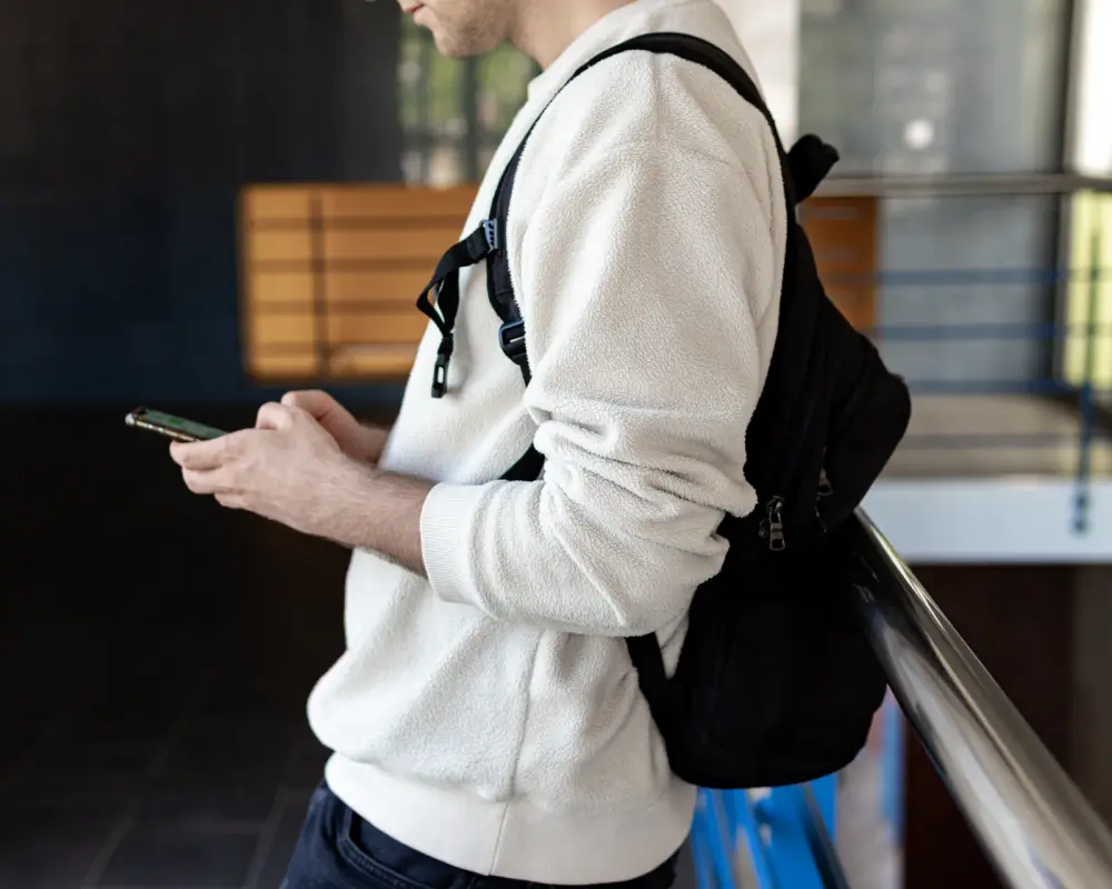 Person in a white jumper and backpack checking a phone by a railing, head out of frame